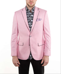 A man wearing a light pink blazer with black piping, a black and white patterned shirt, and black trousers, standing against a plain white background.