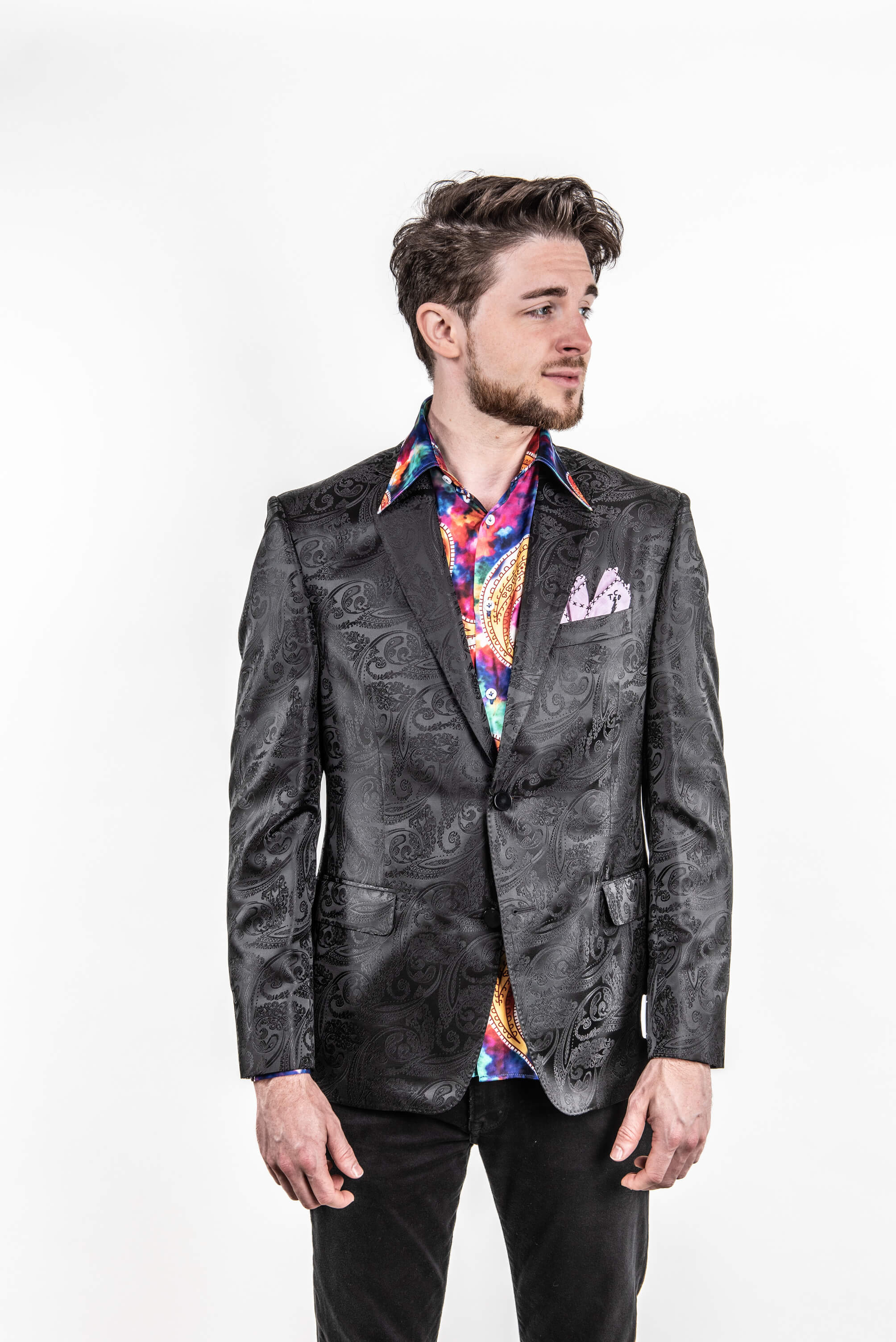 A man wearing a black textured jacquard blazer with a colorful patterned shirt and black trousers, standing against a plain white background.