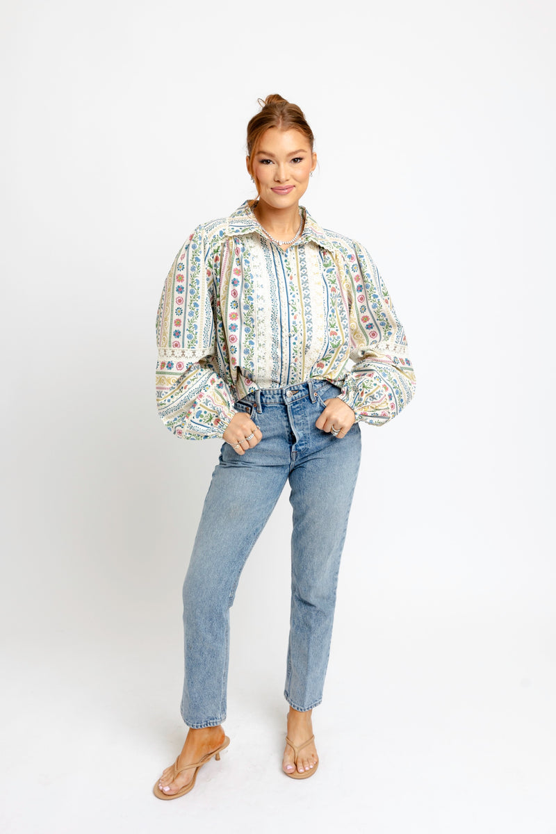 A woman modeling a long-sleeve button-up blouse with a colorful vertical tribal-style pattern, paired with light-wash blue jeans and clear heeled sandals, against a plain white studio background.