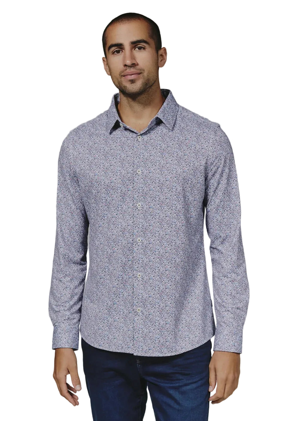 A man wearing a long-sleeve button-up shirt with a dense micro-pattern of small red, blue, and white dots on a light lavender-gray base, paired with dark blue jeans, photographed against a solid gray studio background.