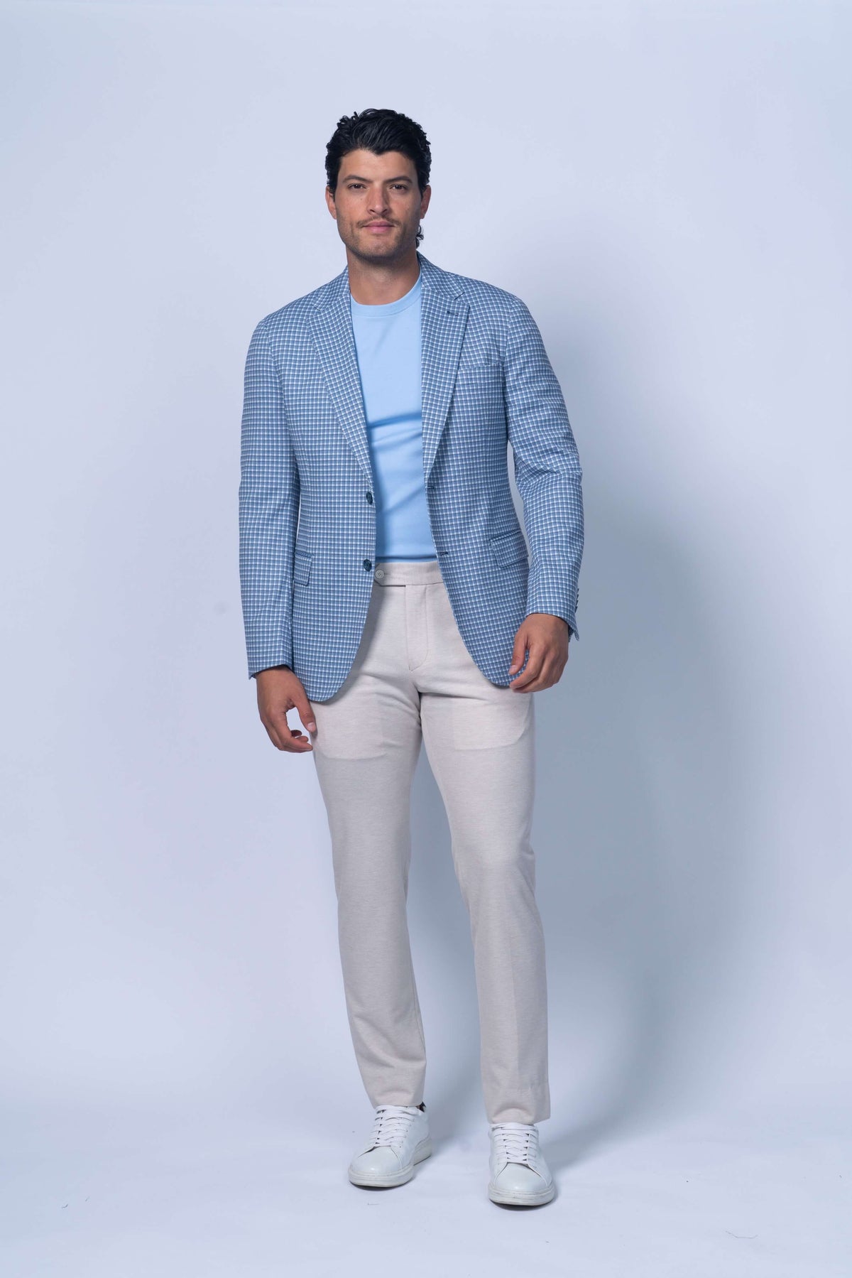 Man wearing a blue checkered blazer, light blue shirt, beige pants, and white sneakers on a white background